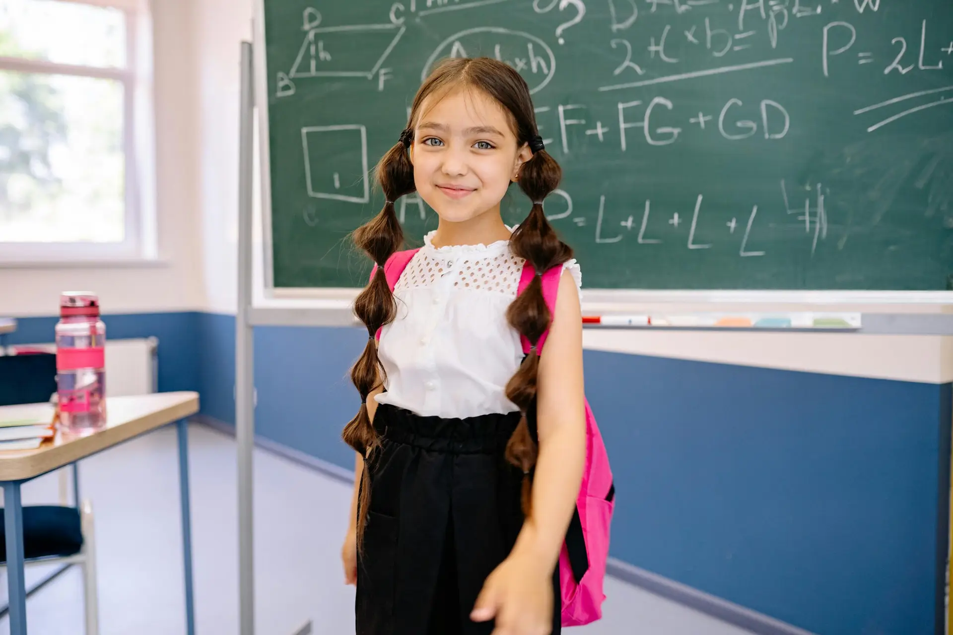 Cute girl with backpack smiling in classroom, standing by the chalkboard with math equations.